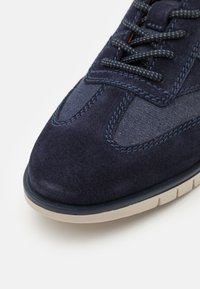 Marinblå sneaker med mocka- och denimpaneler, texturerade snören och en krämfärgad gummisula. Har kontrasterande sömmar och en rundad tå.