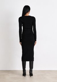 Black ribbed knit dress with long sleeves, fitted silhouette, knee-length, and side vent detail. Paired with tall black heeled boots.