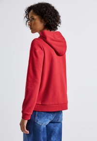 Personne avec des cheveux bouclés courts portant un sweat à capuche rouge et un jean bleu, légèrement tourné vers la gauche devant un fond clair.