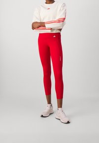 Červené legíny s přiléhavým střihem, doplněné krémovou mikinou s dvěma červenými pruhy na rukávech a logem adidas.