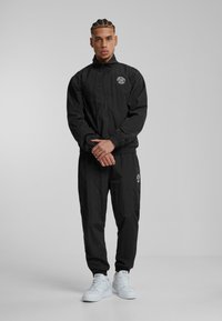 Svart vindtät jacka med framdragkedja, elastiska ärmslut och logopatch. Matchas med byxor i samma tyg och vita sneakers. Slät textur.