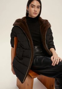 Giacca a vento nera con cappuccio foderato in pelliccia marrone, con una cerniera lucida e design trapuntato. Il modello indossa un dolcevita nero e pantaloni in pelle.