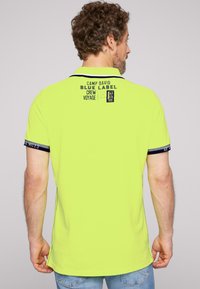 Neongele polo met marineblauwe accenten, voorzien van een logo en tekst op de achterkant, korte mouwen en zijsplitten voor comfort.