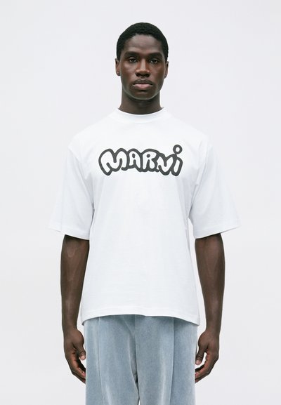 Bărbat îmbrăcat într-un tricou alb supradimensionat cu text negru îndrăzneț "Marni" și pantaloni deschiși, largi, stând în fața unui fundal simplu.