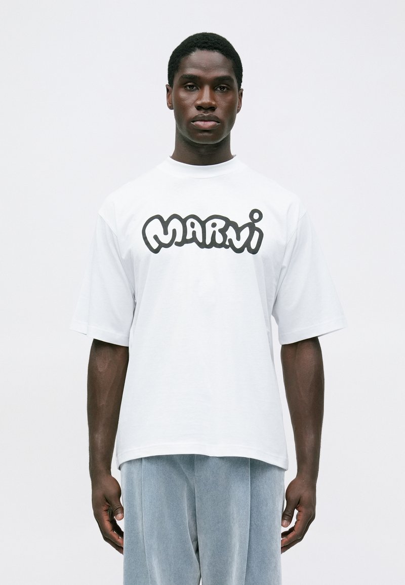 Muž v bílém oversized tričku s výrazným černým nápisem "Marni" a světle modrých volných kalhotách stojící na jednobarevném pozadí.