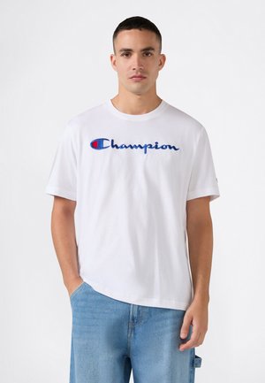 Junger Mann trägt ein weißes Champion-T-Shirt mit blauem Logo und hellblauen Jeans, steht mit einer Hand in der Tasche.