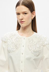 Biela blúzka s prepracovanými kvetinovými zdobenými korálkami na ramene, dlhými rukávmi, zapínaním na gombíky vpredu a hladkou textúrou látky.