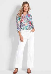 Bloemprint blouse in meerdere kleuren met een V-hals en driekwart mouwen, gecombineerd met witte brede jeans en bruine hakken.