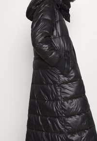 Abrigo largo negro tipo puffer con textura brillante, diseño acolchado, capucha, bolsillos laterales y puños elásticos. Las costuras crean una apariencia ajustada.