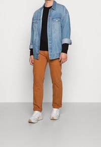Jeansjacke in Hellblau, schwarzer Rippstrickpullover, orange-braune Hose mit Eingrifftaschen und weiße Sneakers mit strukturierten Details.