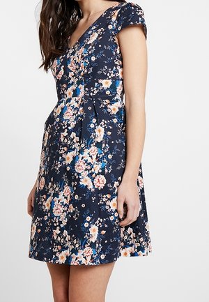 Femme portant une robe bleu marine à manches courtes avec un col en V et un imprimé floral coloré, debout devant un fond blanc uni.