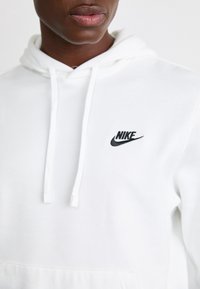 Vit Nike-hoodie med en svart broderad logo. Har en dragsko i huven och ett mjukt, texturerat tyg. Känguruficka på framsidan.