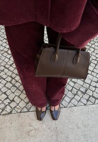 Bolso de cuero marrón con dos asas cortas, combinado con pantalones de pana color burdeos y zapatos de charol marrón, sobre un pavimento estampado.