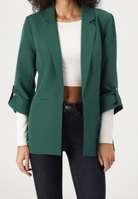 Blazer vert à coupe décontractée, avec des revers crantés et des manches retroussées. Tissu lisse ; porté par-dessus un haut blanc à manches longues et un jean foncé.
