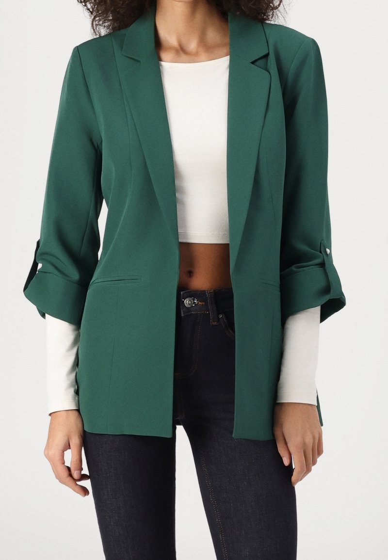 Blazer vert à coupe décontractée, avec des revers crantés et des manches retroussées. Tissu lisse ; porté par-dessus un haut blanc à manches longues et un jean foncé.