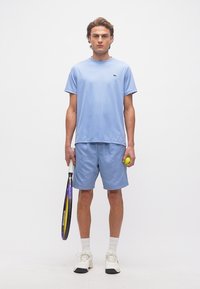 Νεαρός άνδρας με ανοιχτό μπλε αθλητικό ρουχισμό, κρατώντας ρακέτα τένις στο ένα χέρι και μπάλα του τένις στο άλλο, στέκεται μπροστά σε απλό λευκό φόντο.