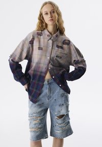 Oversized geruit shirt in lichte tinten met donkerblauwe accenten en versleten denimshorts met grote scheuren en verfsplinterdetails.