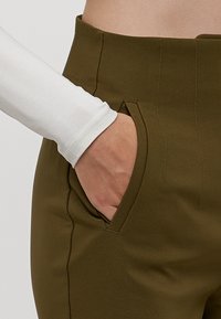 Olive green pants with a smooth, stretchy material and angled side pockets. A white long-sleeve top is partially visible at the cuff.