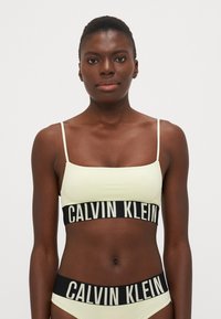 Lys gul bralette med tynne stropper og en svart midjebånd med hvit "CALVIN KLEIN" tekst; jevn tekstur, minimalistisk design.