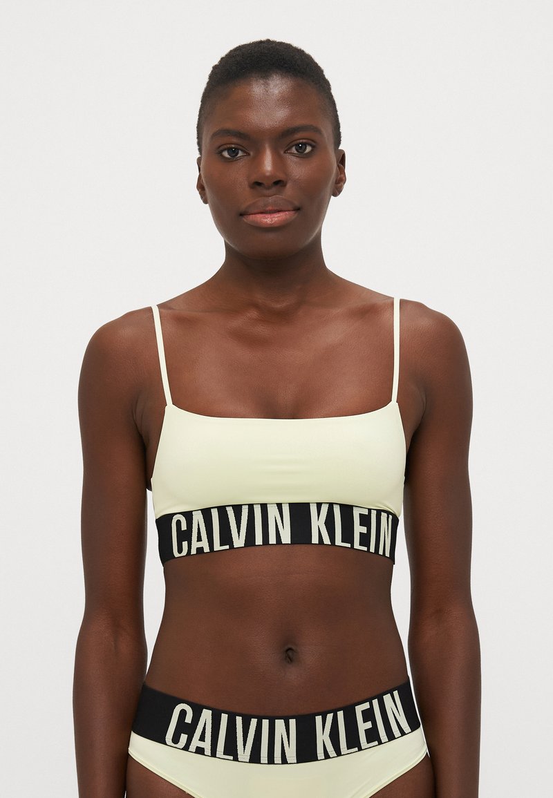 Lys gul bralette med tynne stropper og en svart midjebånd med hvit "CALVIN KLEIN" tekst; jevn tekstur, minimalistisk design.