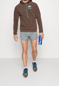 Felpa in pile marrone con dettagli in verde acqua, pantaloni sportivi grigi e scarpe da corsa nere con dettagli blu e gialli, mentre tiene in mano una bottiglia d'acqua blu.