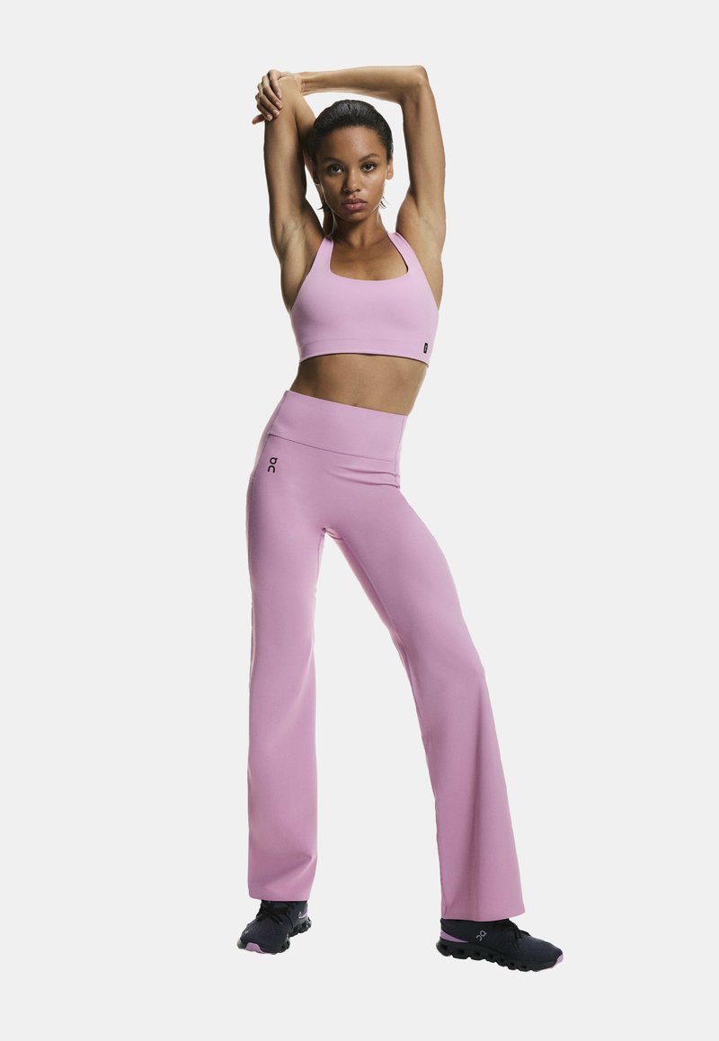 Ružový športový krátky top a legíny s vysokým pásom a rozšírenými nohavicami z pružnej látky. Na páskovej časti výrazné logo, doplnené čiernymi teniskami.