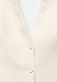 Detailní záběr na smetanovou látku s dvěma odpovídajícími knoflíky potaženými látkou, umístěnými na přední části oděvu s výstřihem do V.