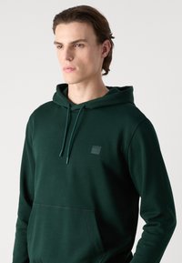 Felpa verde scuro con tasca a marsupio, cappuccio con cordino e un piccolo distintivo nero sul petto. Tessuto morbido con una texture liscia.