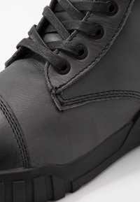 Primer plano de una bota de cuero negro con gruesos cordones negros, costuras visibles y suela de goma robusta sobre un fondo blanco.