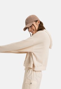 Beige cropped hoodie with a drawstring, soft texture, and wide sleeves, paired with light-colored joggers and a tan cap featuring a logo.