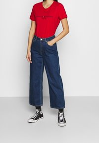 Czerwona bawełniana koszulka z nadrukiem "Tommy Hilfiger", zestawiona z niebieskimi dżinsami o wysokim stanie i szerokimi nogawkami oraz czarnymi trampkami za kostkę.