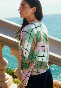 Pull en tricot avec un motif argyle multicolore en vert, lavande, jaune et blanc, entouré de noir. Poignets et ourlet côtelés.