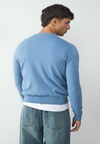 Jasnoniebieski sweter o miękkiej fakturze, dopasowane rękawy i ribbowana talia, nałożony na białą koszulę, noszony z luźno dopasowanymi dżinsami.