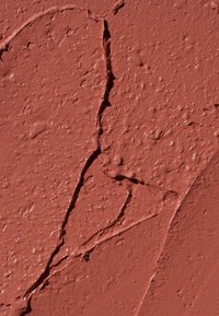 Teksturowana, czerwonobrązowa farba na ścianie z widocznymi pęknięciami i szorstką powierzchnią, ukazująca nieregularne wzory i różne poziomy połysku.