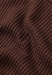 Tissu en maille marron avec un motif côtelé texturé. Présente un petit logo brodé en couleur contrastante. Matière douce et flexible.