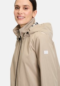 Veste imperméable beige avec fermeture éclair, capuche ajustable et un patch logo blanc sur la manche. Présente un motif à carreaux à l'intérieur de la capuche.