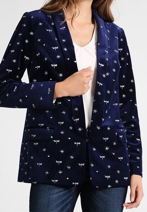 Granatowa marynarka z haftowanymi srebrnymi ważkami i gwiazdami, noszona na białej bluzce i zestawiona z ciemnoniebieskimi dżinsami.