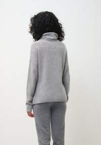 Grijze, lange mouw, coltrui gemaakt van zacht materiaal, gecombineerd met bijpassende grijze loungebroeken, gezien van achteren.