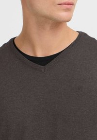 Pulover s V izrezom od tamnosmeđeg pletenog materijala. Ima rebrasti ovratnik i suptilan logotip na prsima. Mekana tekstura, uski kroj.