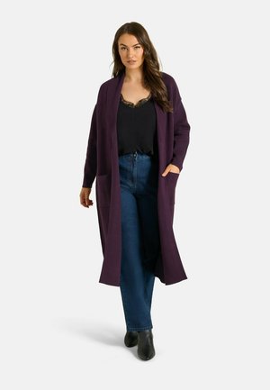 Femme marchant portant un long cardigan violet, un haut noir orné de dentelle, un jean bleu et des bottines noires sur fond blanc.