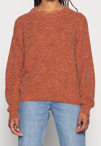 Pull en maille couleur rouille avec un col rond, des manches longues et une coupe décontractée, associé à un jean en denim bleu clair.