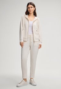 Sweat à capuche beige zippé avec poches avant et cordon de serrage, associé à un pantalon fuselé assorti et des baskets légères. Tissu lisse, design décontracté.