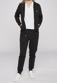 Felpa nera con zip e tasche a marsupio, abbinata a pantaloni della tuta neri con vita elastica e sneakers bianche.