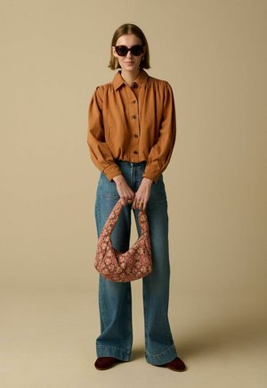 Vrouw met een bruin gebuttond blouse, blauwe spijkerbroek, donkere zonnebril, en een handtas met bloemmotief tegen een effen beige achtergrond.