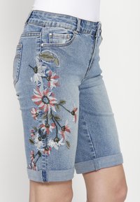 Svetlomodré denim šortky s kvetinovou výšivkou v červenej, bielej a zelenej farbe, s obrúbenými okrajmi a klasickým dizajnom s piatimi vreckami.