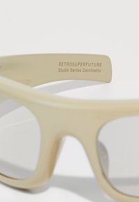 Bež naočale za sunce s glatkim plastičnim okvirom i tamno zatamnjenim lećama. Imaju utisnuti brend na unutrašnjoj strani okvira, "RETROSUPERFUTURE."