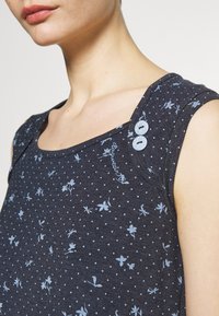 Dark blue sleeveless top with an all-over light blue floral and polka dot pattern. Features two decorative light blue buttons on the shoulder.
