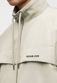 Ελαφριά μπεζ μπουφάν με ψηλό γιακά, φερμουάρ μπροστά και μεταλλικό λογότυπο. Λείο υφή με μινιμαλιστικό σχεδιασμό.