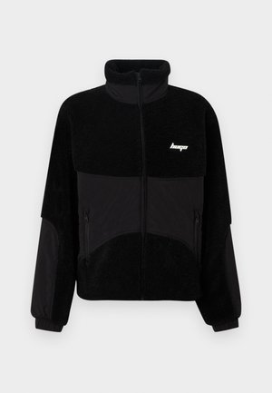 Veste en polaire noire avec un col montant, des panneaux en tissu noir contrastant, des poches latérales zippées et un petit logo sur la poitrine.