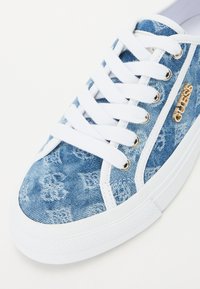 Denim spordijalatsid, millel on sinine pesu, valge kummitald, valged paelad ja kuldne logo detail küljel. Tekstuuriga denim ülemine osa mustritega.